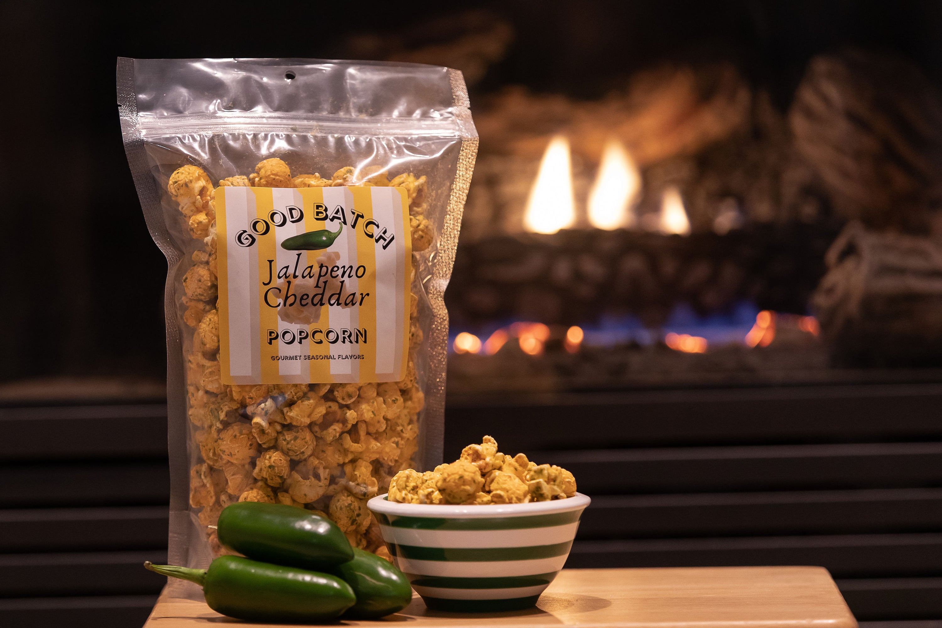 a bag of our gourmet Jalapeno cheddar popcorn pictured in front of a fireplace next to a few fresh jalapenos.